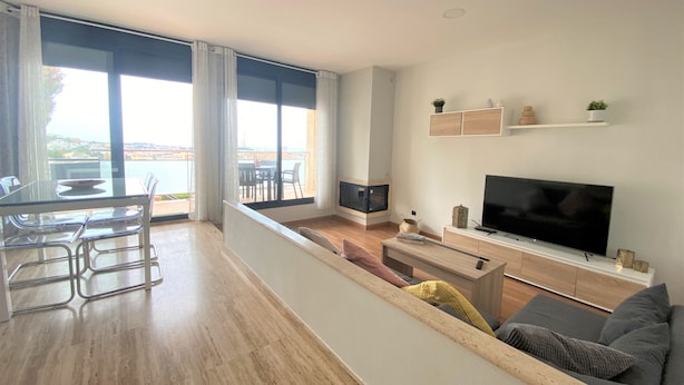 Ein modernes Wohnzimmer mit Flachbildfernseher, einer Couch, einer Essecke mit Tisch und Stühlen und einem großen Fenster mit Meerblick.