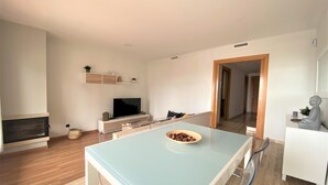 Ein modernes Wohnzimmer mit Fernseher, Holzboden, einem Esstisch mit Obstschale und einer Statue auf einem weißen Schrank.