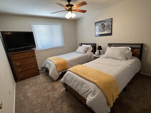 Two single beds with white linens and throw blankets, a dresser, flat-screen TV, and ceiling fan are featured in this room.
