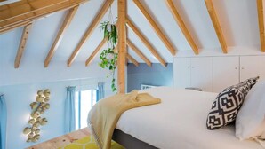 Una camera da letto con soffitto spiovente, travi in legno, un letto con cuscino decorato, una pianta pensile e un addorno a parete.