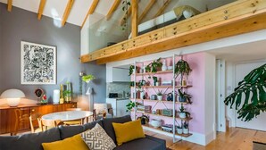 Un soggiorno moderno con un divano nero, travi in legno a soffitto, un'area pranzo e una libreria piena di piante.
