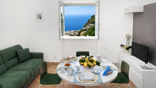 A modern room with a dining table set for two, a green sofa, a TV, and a window with a coastal view.