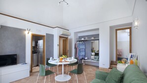 A modern living room with a green sofa, a dining table with chairs, a TV, and a kitchen area with a refrigerator and a sink.