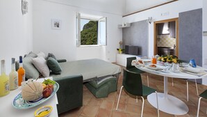 A compact room with a bed, a dining table with chairs, a kitchen area, and a window with a view of greenery.