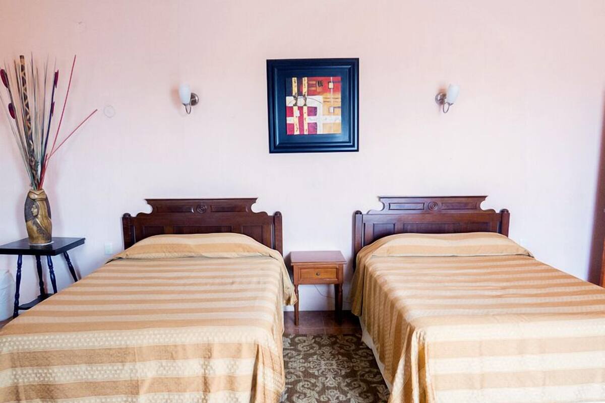 A room with two beds, a small table, a vase with dried plants, and a framed artwork on the wall.