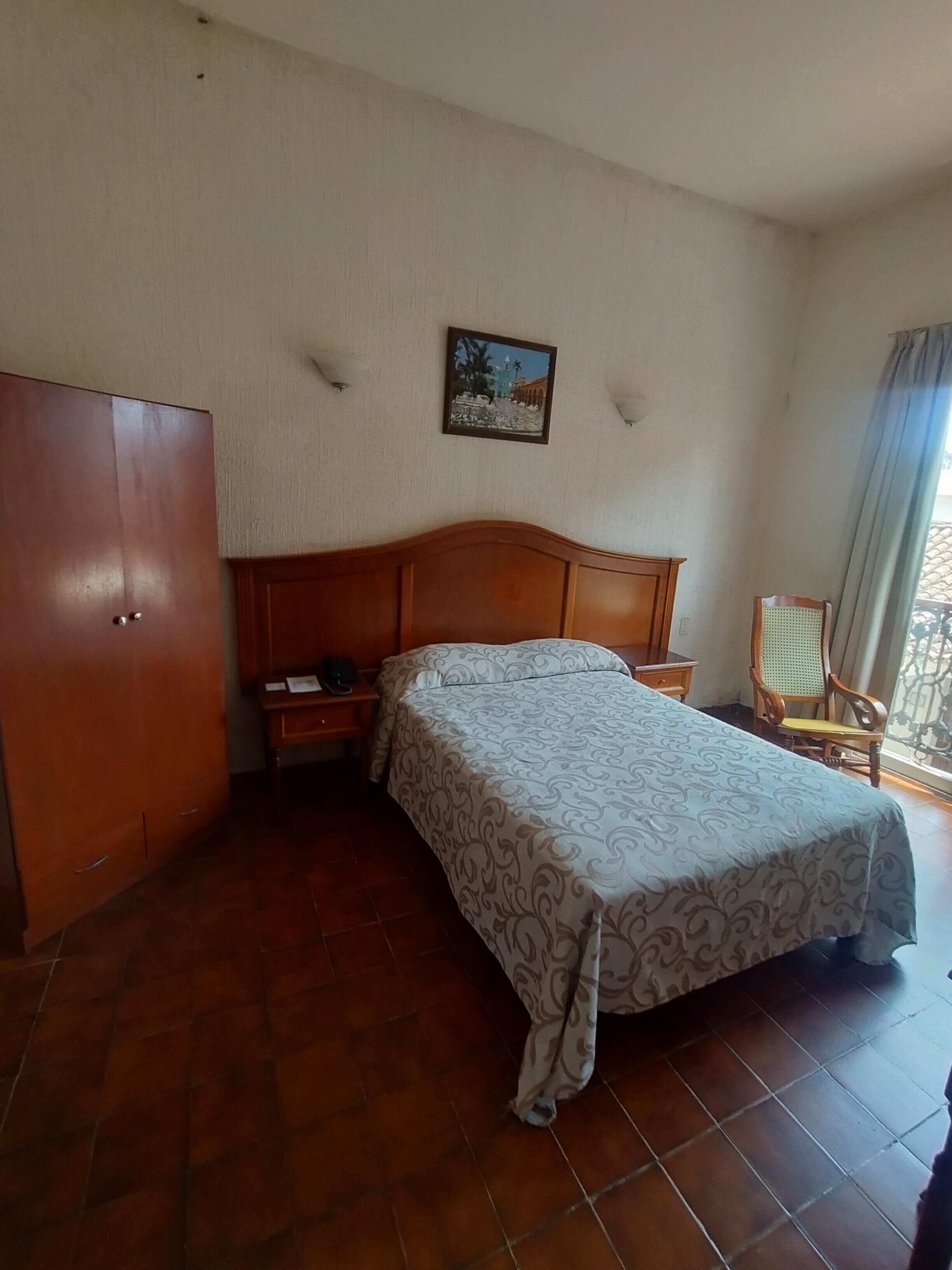 A bedroom with a wooden bed, a nightstand, a wardrobe, a picture on the wall, and a chair by the window.