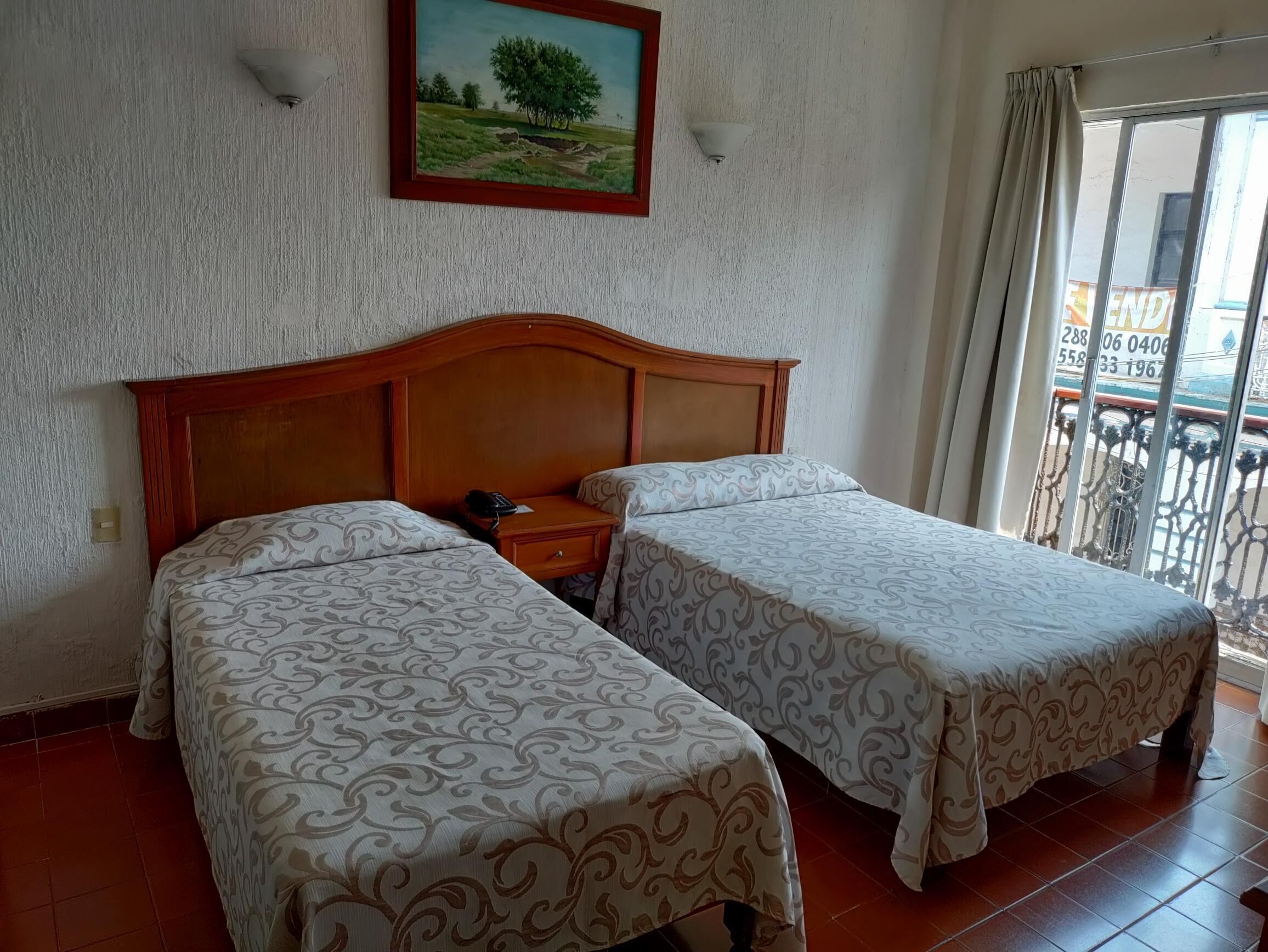 Ein Hotelzimmer mit zwei Einzelbetten, einem Holzkopfende, einem Nachttisch, einem Bild an der Wand und einem Balkon mit Blick.