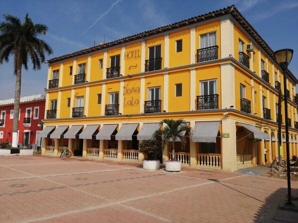 Un hotel amarillo con balcones y toldos, situado en una plaza con palmeras y un edificio rojo cercano.