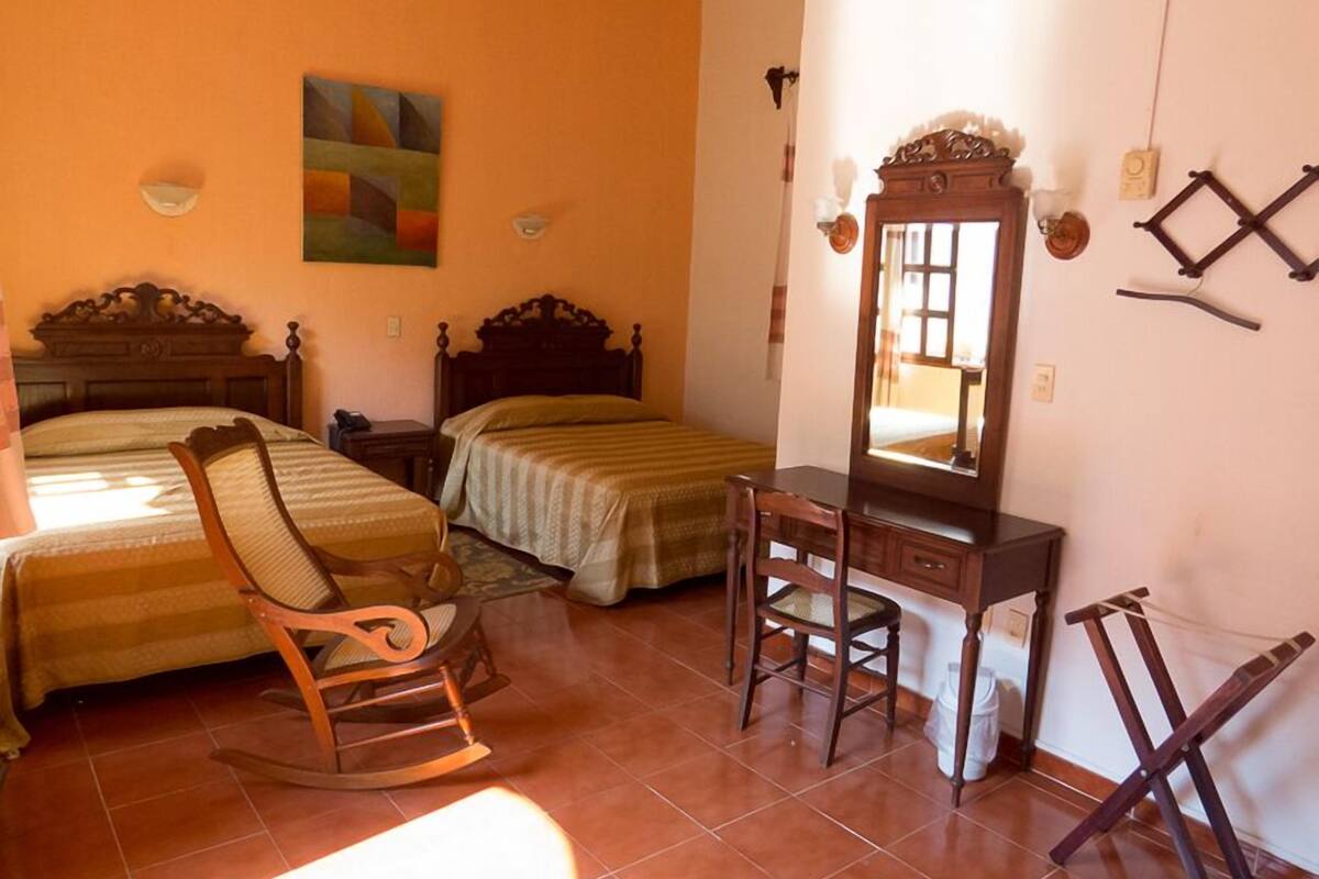 A bedroom with two beds, a wooden rocking chair, a desk with a mirror, and a painting on the wall.