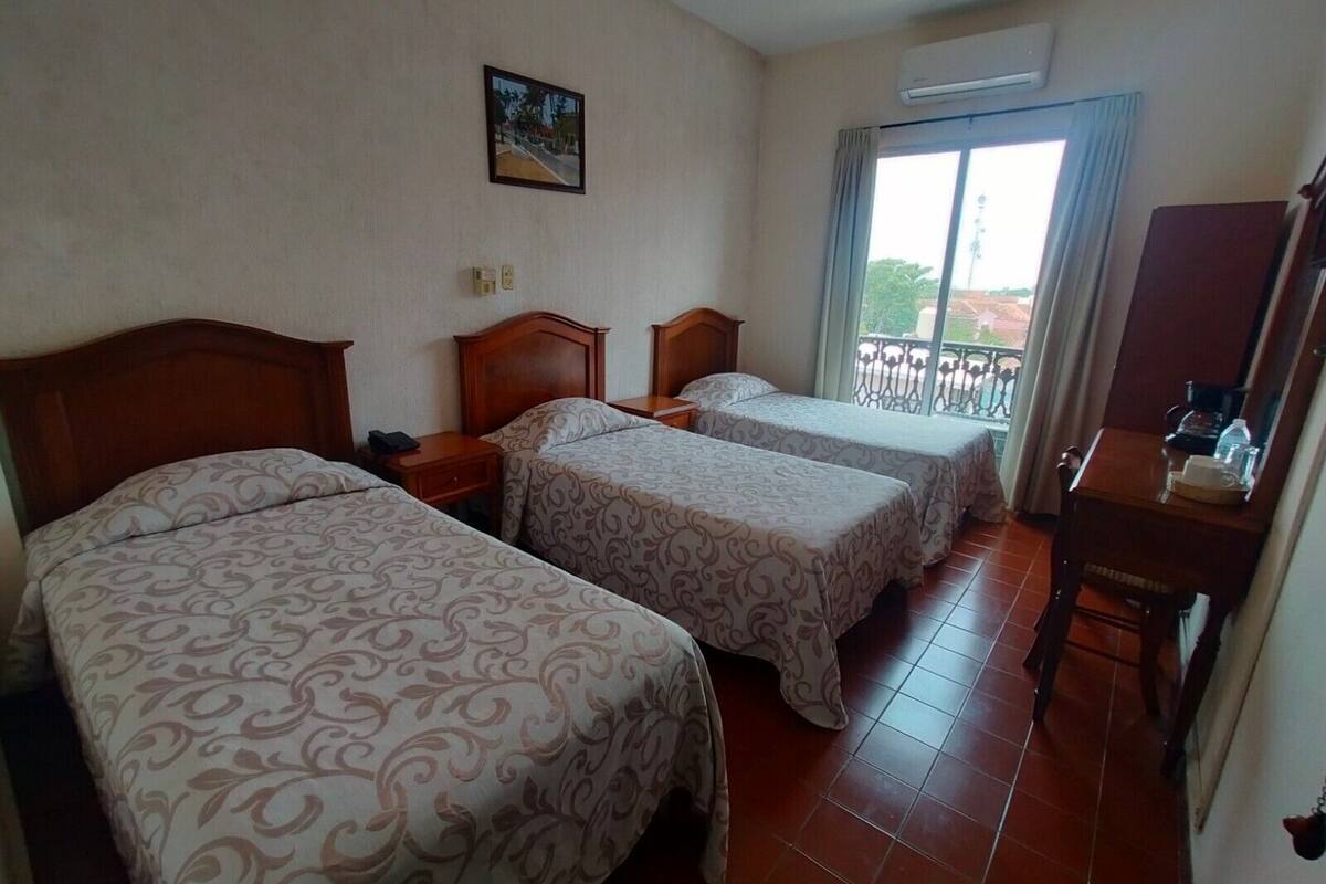 A hotel room with two beds, a wooden nightstand, a desk with a chair, and a balcony with a railing.