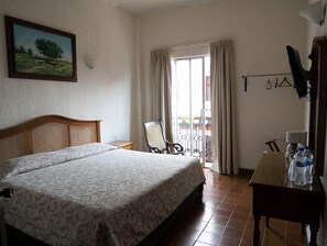 A hotel room with a bed, a chair, a table with water bottles, a painting on the wall, and a balcony with curtains.