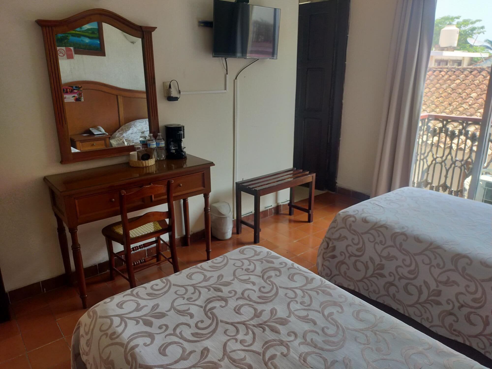 Habitación de hotel con dos camas, un escritorio con silla, un espejo, un televisor montado en la pared y una ventana con vista a edificios.