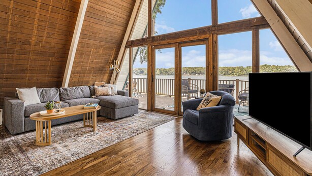 A spacious loft with a high ceiling, large windows, a cozy seating area with a sofa and armchair, and a flat-screen TV on a wooden stand. - Lakefront A-Frame: Private Beach, Fire Pit, Kayaks, Paddleboards, & Boat Slip (Decatur)