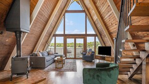 A spacious loft with a high ceiling, wooden beams, a large window, a fireplace, a sofa, a chair, and a staircase. - Lakefront A-Frame: Private Beach, Fire Pit, Kayaks, Paddleboards, & Boat Slip (Decatur)