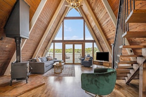 A spacious loft with a high ceiling, wooden beams, a large window, a fireplace, a sofa, a chair, and a staircase.