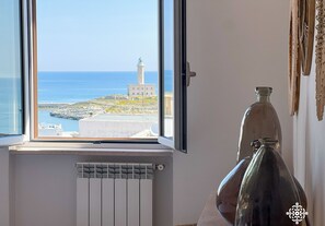 Quarto com grande janela que oferece vista para a costa, incluindo um farol e terreno rochoso. Um radiador é visível abaixo da janela, e um vaso de vidro decorativo está sobre uma mesa à direita.
