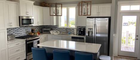 Una cocina moderna con armarios blancos, una isla grande con taburetes de bar, electrodomésticos de acero inoxidable y vistas al exterior.