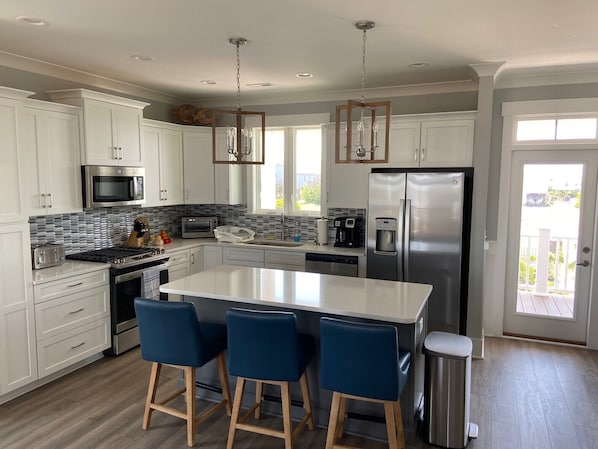 A modern kitchen with white cabinets, a large island with bar stools, stainless steel appliances, and a view to the outside.