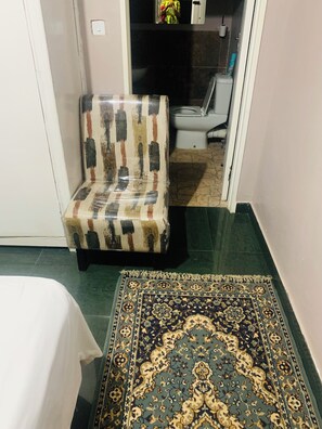 A room with a patterned armchair, a large decorative rug, and a toilet visible through an open door.