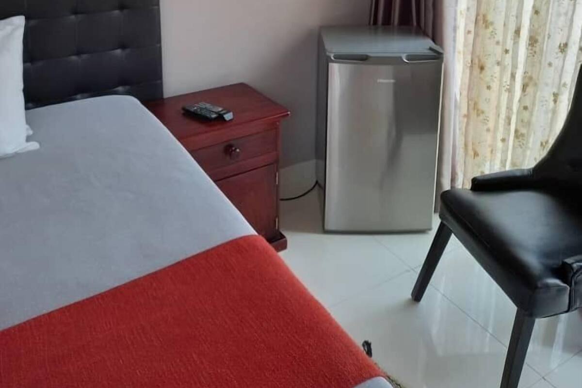 Habitación de hotel con cama, un escritorio pequeño con lámpara, una silla, refrigerador y ventana con cortinas.