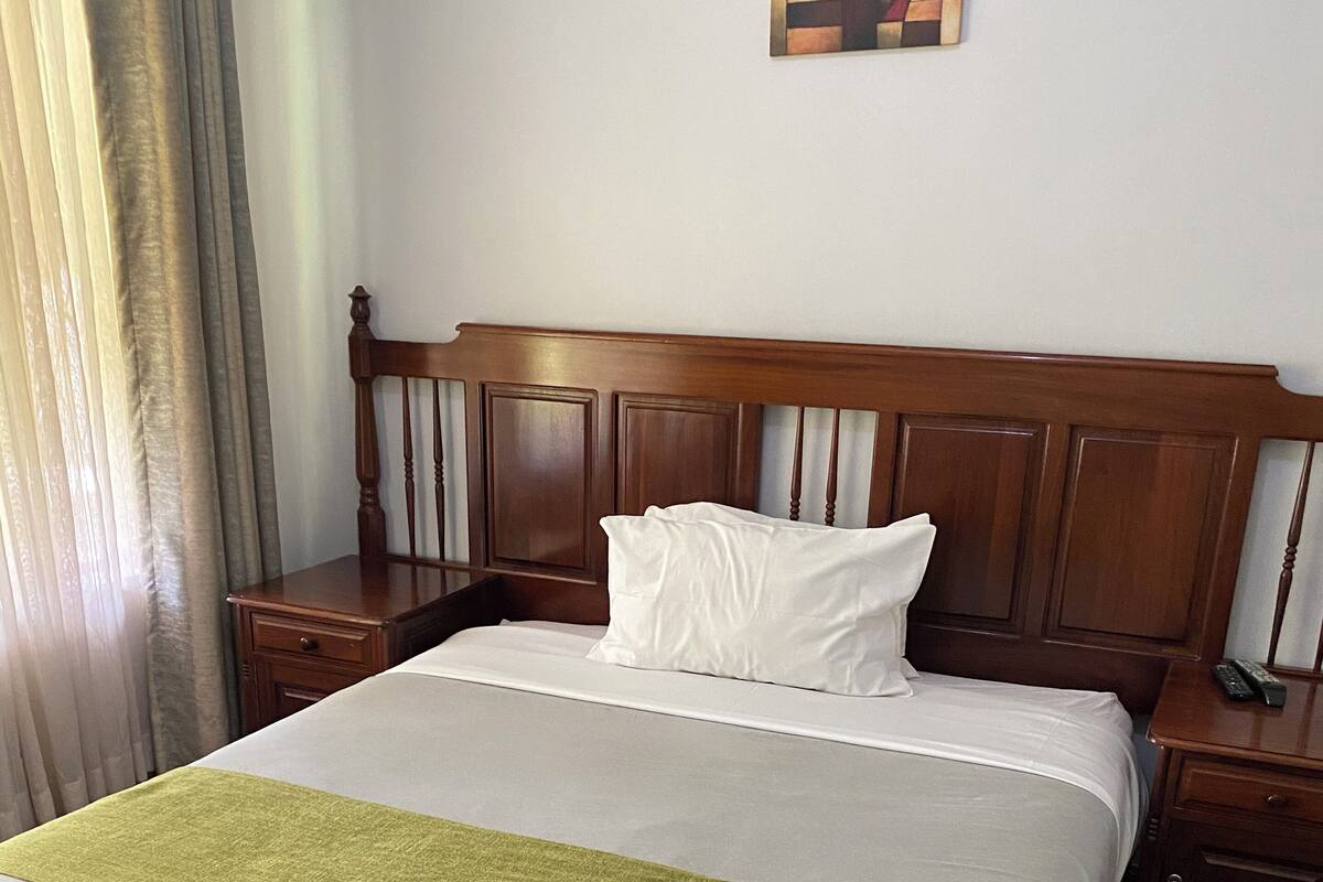 Habitación de hotel con una cama bien hecha, cabecera de madera, mesita de noche con un control remoto y un cuadro en la pared.