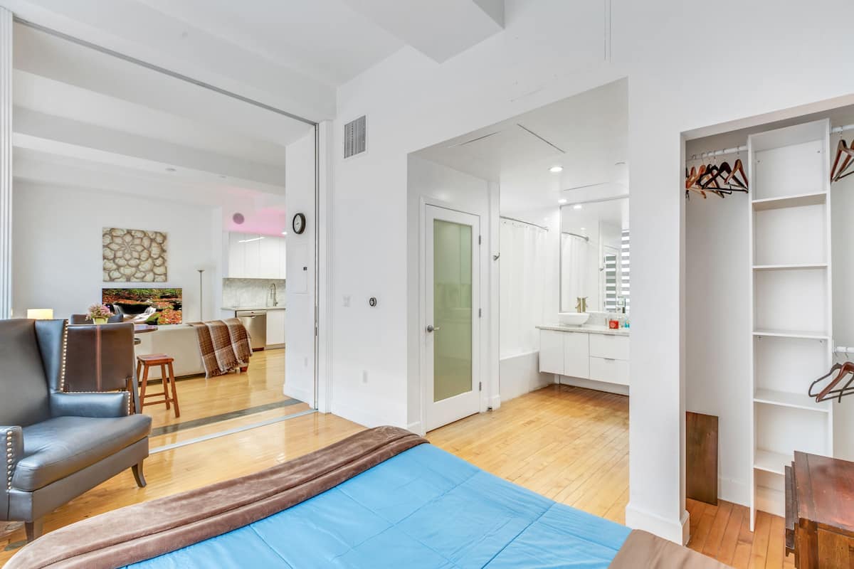 Une chambre d’hôtel moderne comprenant un lit, un coin salon avec un fauteuil, une table à manger et un coin cuisine visible à travers une porte ouverte.