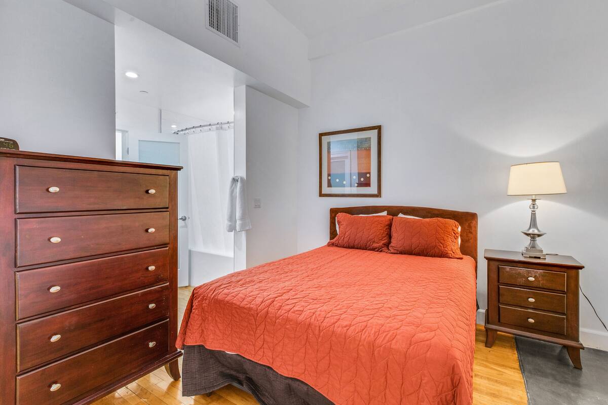 Une chambre avec une commode en bois, un lit recouvert d’une courtepointe orange, une table de chevet avec une lampe et un tableau encadré au mur.