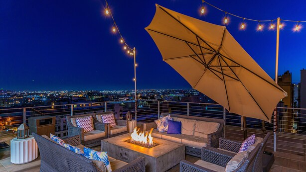 A rooftop terrace with wicker furniture, a fire pit, string lights, and a view of the cityscape at night.
