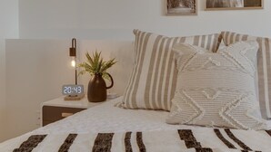 Un lit bien fait, agrémenté d’oreillers à rayures et d’un plaid tricoté, une table de chevet avec une horloge numérique et une petite plante, et un tableau encadré au mur.