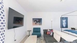 Ein modernes Wohnzimmer mit Flachbildfernseher, einem türkisfarbenen Sessel, einem grauen Sofa und einem gemusterten Teppich.