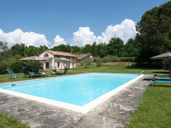A rectangular swimming pool with a stone border, surrounded by a grassy area with lounge chairs and umbrellas.