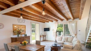 A spacious living area with a wooden ceiling, a dining table with chairs, a sofa, and a TV mounted on the wall.