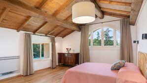 A bedroom with a wooden ceiling, a bed with a pink bedspread, a bedside table with a vase, and a window with curtains.