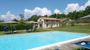 A swimming pool with lounge chairs and umbrellas, surrounded by a grassy area and a building with a tiled roof.