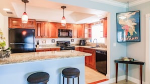 A modern kitchen with wooden cabinets, a black refrigerator, a microwave, and a granite countertop. There are two bar stools and a small table with a picture frame. - Zen Beach Villa w/ Pool & Garden Views, BBQ (Naples)