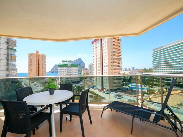 A balcony with a table and chairs, a glass railing, and a view of buildings and a mountain.