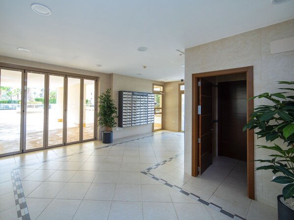 Un hall d’entrée spacieux avec un sol carrelé, une plante en pot, un écran fixé au mur et une porte d’ascenseur en bois.