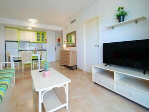 A modern living room with a flat-screen TV, a dining area with a table and chairs, and a kitchen with green cabinets.