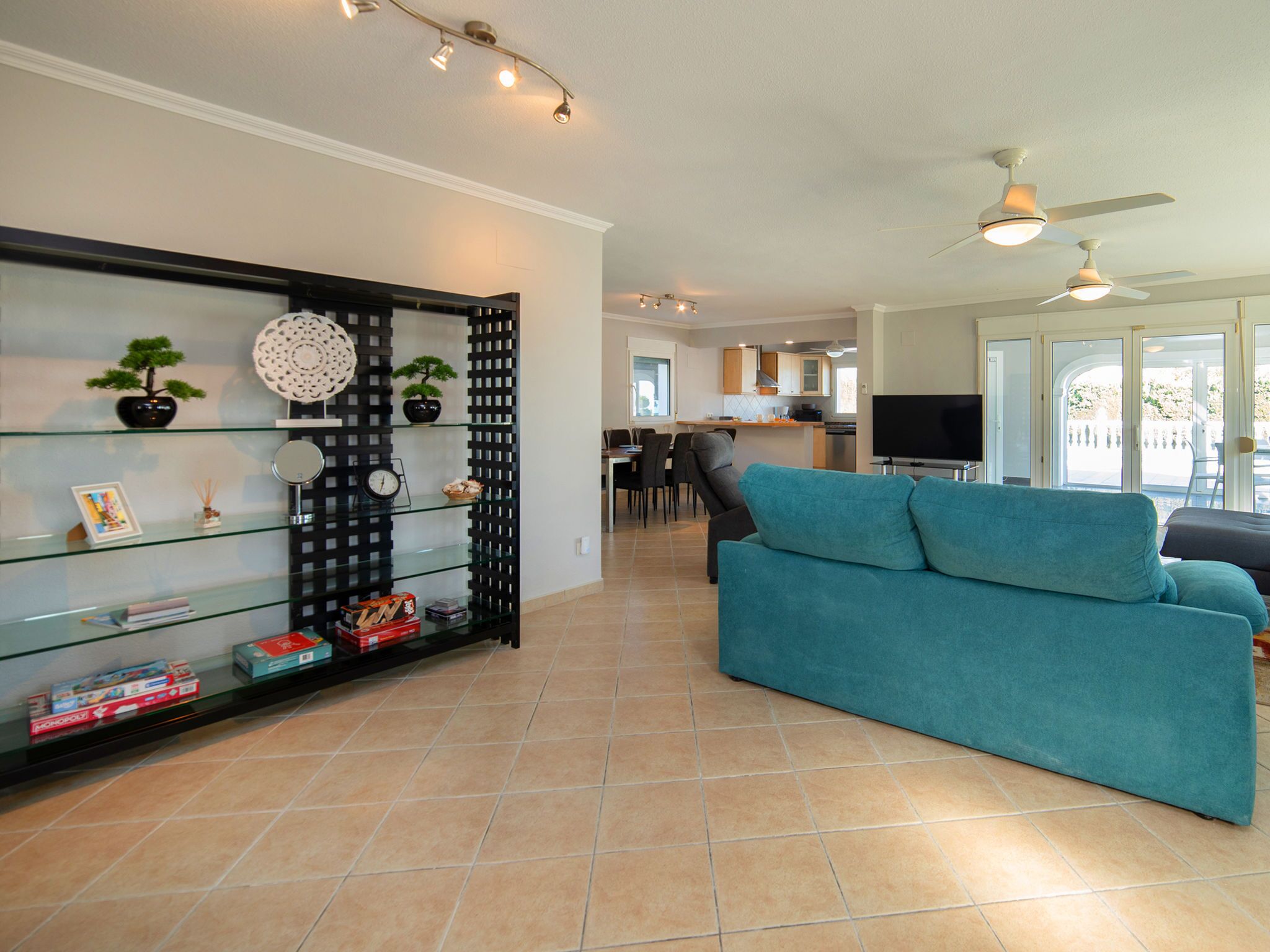 A modern living room with a teal sofa, a glass shelving unit with decorative items, a dining area, and a kitchen in the background.