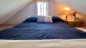 Ein Schlafzimmer mit schrägem Dach, einem Bett mit dunkelblauer Bettdecke, einem Nachttisch mit Lampe und Pflanze sowie einem Fenster mit Blick nach außen.
