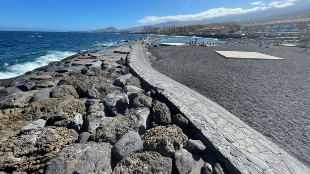 A rocky coastal walkway leading to a pebble beach with a clear view of the ocean and distant buildings. - Arenitas Beach.  An oasis by the sea (CN)