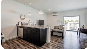A modern kitchen with dark cabinets, a central island, pendant lighting, and a dining area with a sofa and table.