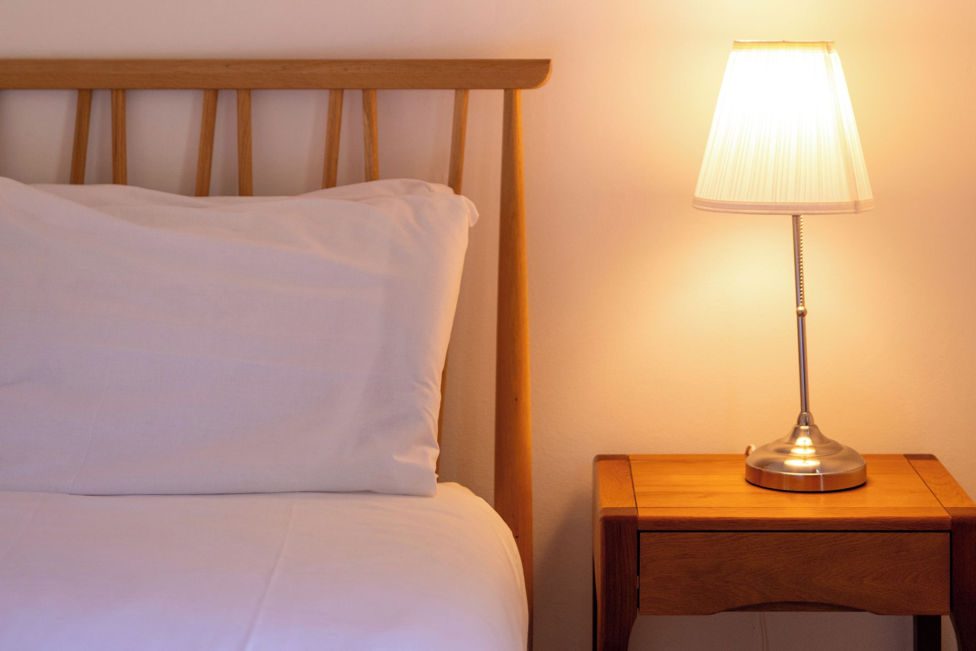 A bed with white bedding and a wooden headboard, a bedside table with a lamp, and a wall with a light switch.
