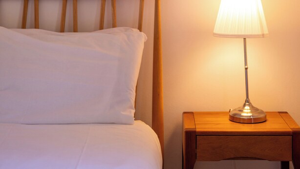Ein Bett mit weißer Bettwäsche und einem Holzkopfteil, ein Nachttisch mit Lampe sowie eine Wand mit Lichtschalter.