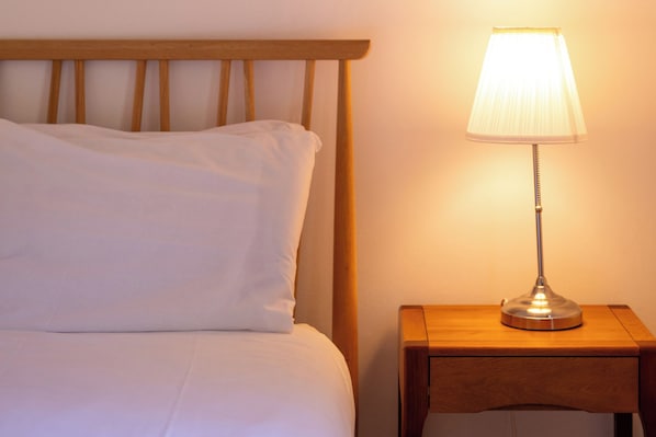 A bed with white bedding and a wooden headboard, a bedside table with a lamp, and a wall with a light switch.