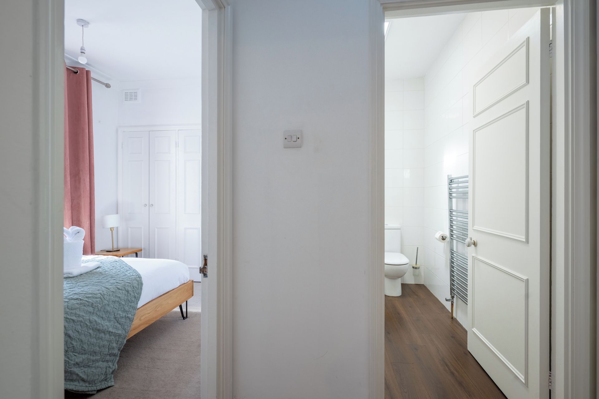 Una habitación de hotel con cama, mesita de noche, lámpara y baño equipado con inodoro y toallero.