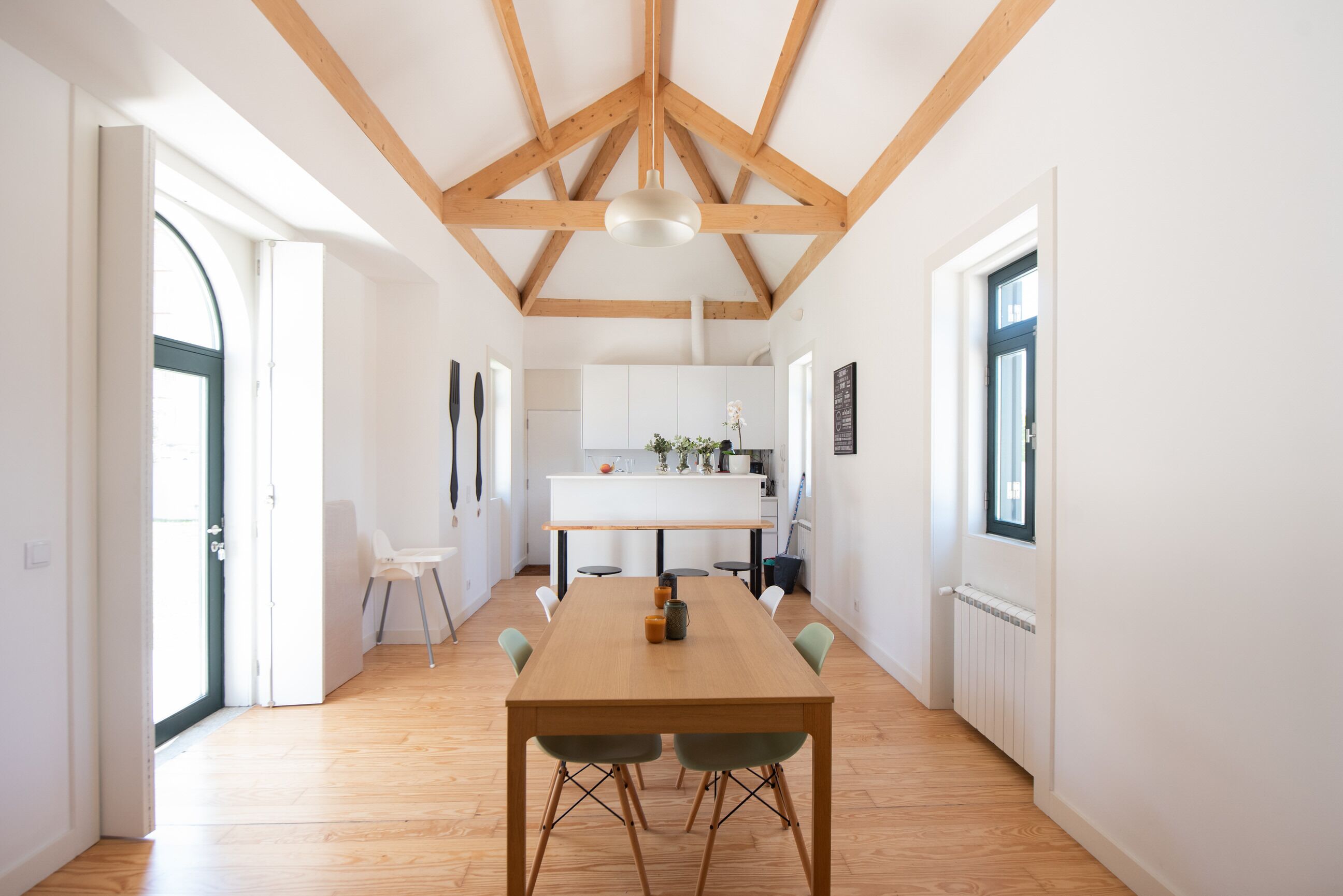 A spacious dining area with a wooden table and chairs, a kitchen in the background, and a high ceiling with exposed beams.