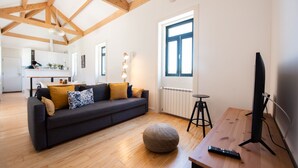 A modern living room with a dark sofa, wooden flooring, a round ottoman, a TV on a wooden stand, and a kitchen in the background. - Casa São João a 400 Metros da Praia com Jardim (Arcozelo)