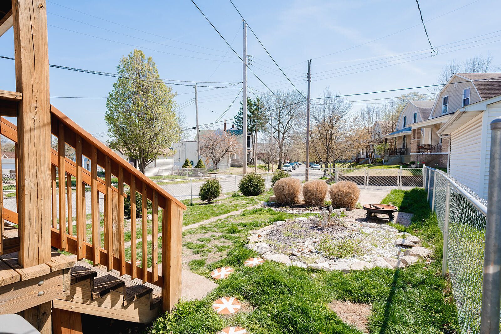A residential backyard with a wooden deck, a stone pathway, and a small garden area with shrubs and a wheelbarrow.