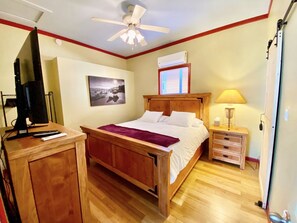 A bedroom with a wooden bed, a desk with a computer, a ceiling fan, a window with blinds, and a nightstand with a lamp.
